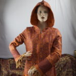 In einem lebendigen Kleiderschrank erzählt der Mantel seine Geschichte: "Ich bin jugendlich, frech und bereit, die Welt zu erobern. Mit floralen und Paisley Mustern, in gedecktem Orange und harmonischem Creme Orange, bin ich ein lebendiges Unikat mit handwerklichen Narben. Nicht nur für den Kleiderschrank gemacht, sondern für Straßen und unvergessliche Abenteuer. Mein Stoff trägt Leidenschaft und Freiheit. Ich bin der freigeistige Träumer, bereit, die Welt mit Stil zu erobern."