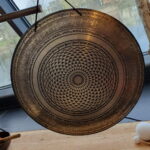 die Schönheit des Klangs mit unserem prachtvollen Bronce-Gong. Ein Kunstwerk aus der Mitte strömender Dreiecke, die sich spiralförmig nach außen verteilen und in kunstvoll verzierten Ringen enden, auf denen ein heiliges Mantra in Sanskrit ruht. Über allem thronen faszinierende Verzierungen, die diesem majestätischen Instrument eine Aura der Erhabenheit verleihen.