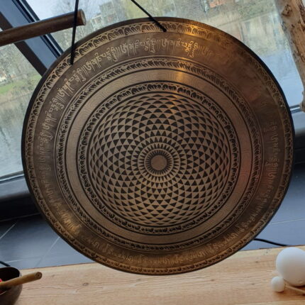 die Schönheit des Klangs mit unserem prachtvollen Bronce-Gong. Ein Kunstwerk aus der Mitte strömender Dreiecke, die sich spiralförmig nach außen verteilen und in kunstvoll verzierten Ringen enden, auf denen ein heiliges Mantra in Sanskrit ruht. Über allem thronen faszinierende Verzierungen, die diesem majestätischen Instrument eine Aura der Erhabenheit verleihen.